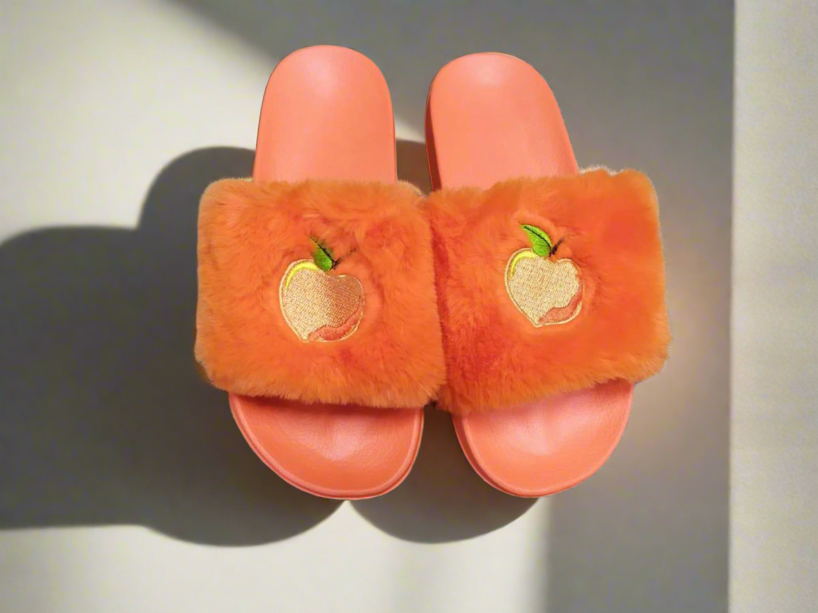 A pair of orange fuzzy slide slippers with an embroidered peach on the strap, placed on a wooden floor.