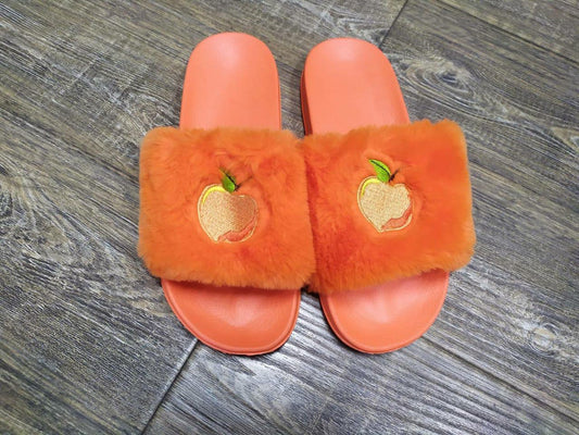 A pair of orange fuzzy slide slippers with an embroidered peach on the strap, placed on a wooden floor.